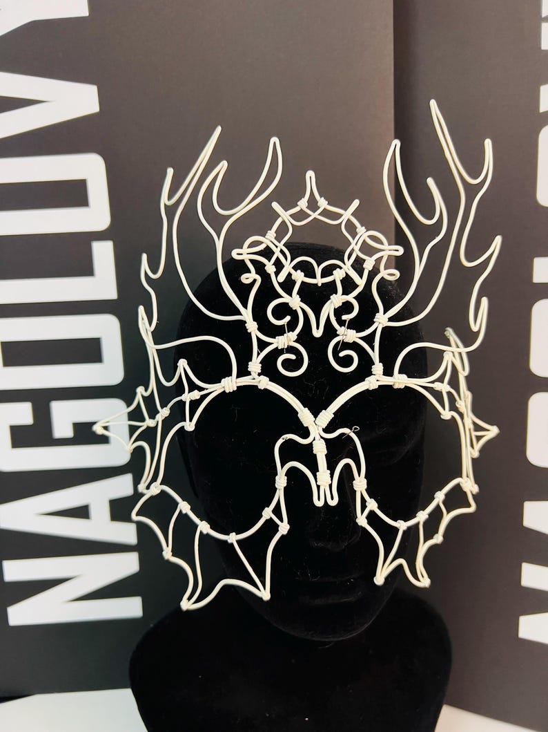 May include: A white wire crown with an intricate design featuring antler-like shapes and webbed patterns. The crown is displayed on a black mannequin head against a black background with white text.