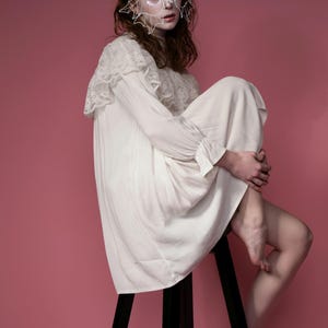 May include: A white, long-sleeved dress with lace detailing on the shoulders and cuffs. The person is wearing a white, wire-frame mask and is seated on a black stool against a pink background.