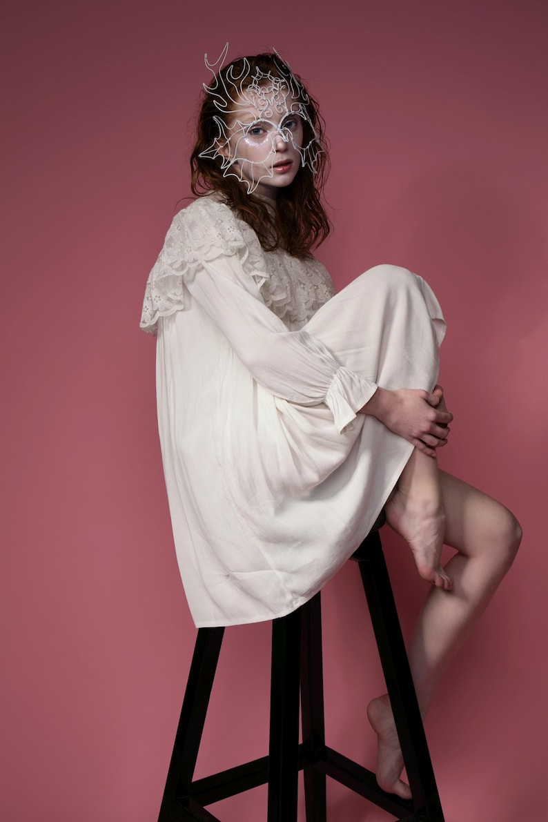 May include: A white, long-sleeved dress with lace detailing on the shoulders and cuffs. The person is wearing a white, wire-frame mask and is seated on a black stool against a pink background.
