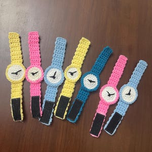 May include: A collection of seven colorful, crocheted toy watches. Each watch features a round face with a black clock hand and a Velcro strap. The watches are in various colors, including yellow, pink, blue, and teal.