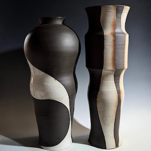 Puede incluir: Dos jarrones altos de cerámica. El jarrón de la izquierda es negro con un diseño blanco moteado y curvo. El jarrón de la derecha es negro y blanco con rayas verticales. Ambos jarrones tienen un acabado mate.