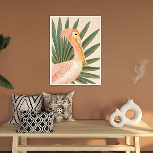 May include: A pink pelican with a long beak is depicted in a watercolor style against a background of green palm leaves. The artwork is framed and displayed on a wall.