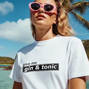 Op de afbeelding: Wit T-shirt met de tekst "give me gin & tonic" in een zwarte rechthoek. De foto is buiten genomen met een tropische achtergrond, inclusief een palmboom en blauwe lucht. De persoon draagt een roze zonnebril.