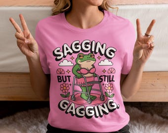 Op de afbeelding: Roze T-shirt met de tekst "SAGGIN' BUT STILL GAGGIN'". De afbeelding toont een groene kikker met een roze bh en rok, die een rollator gebruikt. Het ontwerp bevat wolken, sterren en bloemen.