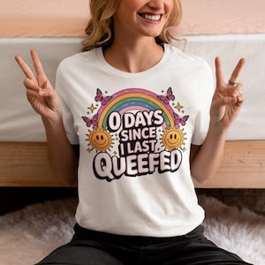 May include: White t-shirt with a colorful graphic design. The design includes a rainbow, butterflies, smiling suns, and the text "0 Days Since I Last Queefed."