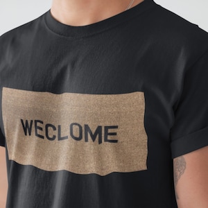 May include: A black t-shirt with a brown rectangle featuring the text "WECLOME" in black.
