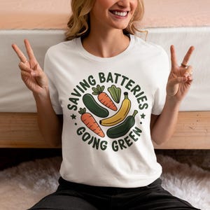 May include: White t-shirt with a circular graphic featuring vegetables and the text "SAVING BATTERIES GOING GREEN". The design includes carrots, a cucumber, a banana, and a pepper. The shirt is worn by a person making a peace sign.
