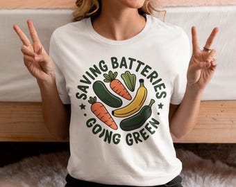 May include: White t-shirt with a circular graphic featuring vegetables and the text "SAVING BATTERIES GOING GREEN". The design includes carrots, a cucumber, a banana, and a pepper. The shirt is worn by a person making a peace sign.