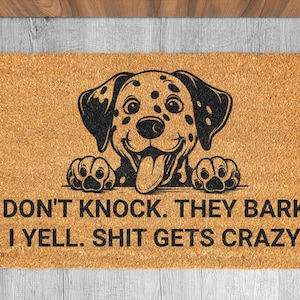 Op de afbeelding: Rechthoekige kokosdeurmat met een zwart-wit Dalmatische hond afbeelding. De tekst "DON'T KNOCK. THEY BARK. I YELL. SHIT GETS CRAZY" is in zwart gedrukt. De mat is bruin en geschikt voor een deuropening.