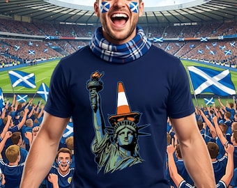 May include: A navy blue t-shirt featuring a graphic of the Statue of Liberty holding a torch and wearing a traffic cone hat. The image is set against a stadium backdrop with Scottish flags and fans.