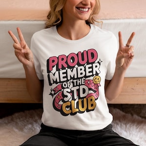 May include: White t-shirt with the text "PROUD MEMBER OF THE STD CLUB" in pink, black, and yellow. The design includes a smiley face and ribbon accents. The shirt is a crew neck.
