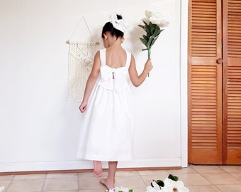 Op de afbeelding: Een jong persoon draagt een witte jurk met een grote strik op de rug en een bijpassende scrunchie. De persoon houdt witte bloemen vast. Een macramé wandkleed en een houten deur zijn op de achtergrond te zien.