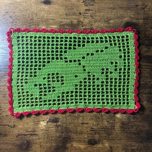 May include: A rectangular crochet doily featuring a light green grid pattern with a central design of two stylized elephants. The piece is finished with a scalloped red border that frames the entire perimeter of the green crocheted fabric.