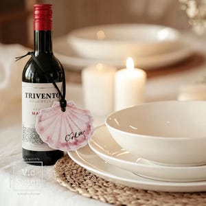May include: A bottle of red wine with a red cap and a black label. A pink seashell-shaped tag with the name "Orla" hangs from the bottle. White plates, bowls, and candles are in the background. The scene is set on a woven placemat.