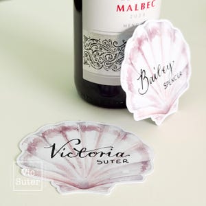 May include: Two seashell-shaped place cards with names, "Bailey Spencer" and "Victoria Suter", in elegant script. The cards have a watercolour effect in shades of pink and white. A wine bottle is partially visible in the background.