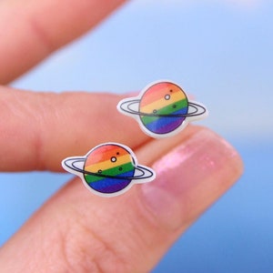 May include: A pair of rainbow pride planet earrings with a white background. The planets have a ring around them and are rainbow colored.