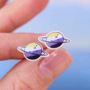 Puede incluir: Un par de pequeños pendientes de botón blancos con un diseño de planeta amarillo, morado y blanco. Los planetas tienen un anillo negro delgado alrededor.