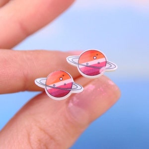 May include: A pair of stud earrings featuring a white planet with a pink, orange, and red gradient design. The planet has a thin ring around it.