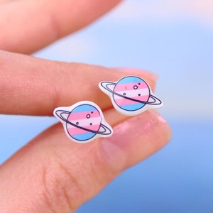 May include: A pair of white stud earrings with a blue, pink, and light blue planet design. The planet has a ring around it and a few small white dots.