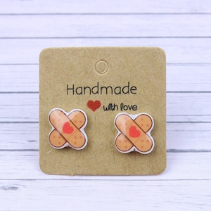 May include: A pair of small, brown and white earrings shaped like plasters with a red heart in the centre. The earrings are displayed on a brown card with the text "Handmade with love".