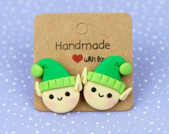 May include: A pair of handmade elf-themed earrings. Each earring features a smiling elf face with a green hat and a yellow brim. The earrings are attached to a brown card with the text "Handmade with love".