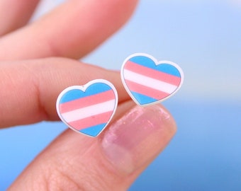 May include: A pair of heart-shaped earrings with the transgender pride flag colors: light blue, pink, and white.