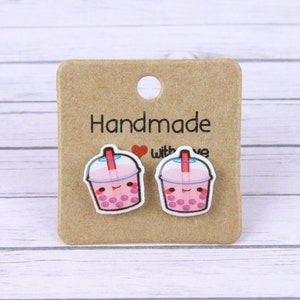 Op de afbeelding: Een paar roze en witte bubble tea oorbellen met een rietje en een glimlachend gezicht. De oorbellen zijn op een bruine kaart met de tekst "Handmade with Love".