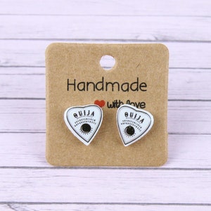 May include: A pair of handmade heart-shaped earrings with a white background and black "OUIJA" lettering. The earrings are on a brown card with the text "Handmade with love".