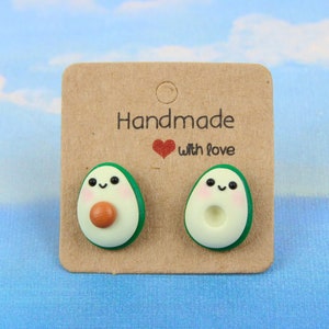 Può includere: Un paio di orecchini a forma di avocado con un viso carino e un seme marrone. Gli orecchini sono su una carta marrone con il testo "Handmade with love".