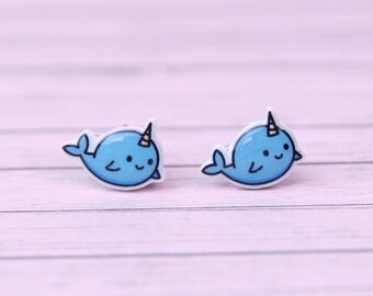 May include: A pair of blue narwhal stud earrings with white outlines and a yellow horn. The earrings are on a light pink wooden surface.