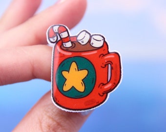 May include: A red mug with a green circle and a yellow star, filled with brown liquid and topped with white marshmallows and a red and white candy cane.