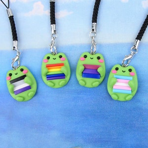 Może przedstawiać: Pięć zielonych breloczków do telefonu w kształcie żaby z różnymi flagami dumy. Flagi to tęcza, biseksualna, transpłciowa, aseksualna i niebinarna.