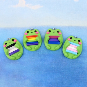 May include: Four green frog figurines holding different pride flags. The flags are rainbow, bisexual, asexual, and transgender.