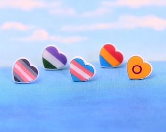 May include: Five heart-shaped pins with different pride flag designs. The pins are white with colorful stripes and symbols.