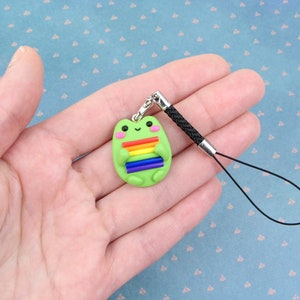 K&ouml;nnte beinhalten: Ein gr&uuml;ner Frosch-Anh&auml;nger mit einer Regenbogenflagge an einem schwarzen Handyband.