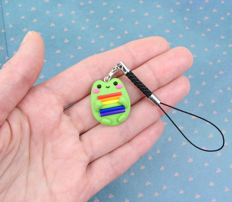 K&ouml;nnte beinhalten: Ein gr&uuml;ner Frosch-Anh&auml;nger mit einer Regenbogenflagge an einem schwarzen Handyband.