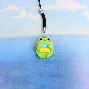May include: A green frog charm with a yellow and blue stripe on its belly, holding a yellow and blue striped rectangle. The charm is attached to a black string.