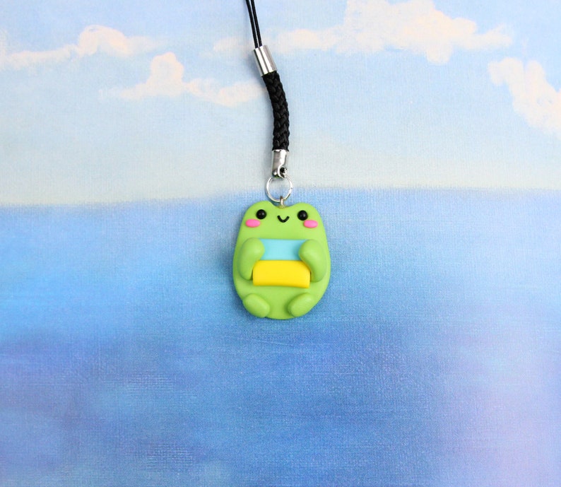 May include: A green frog charm with a yellow and blue stripe on its belly, holding a yellow and blue striped rectangle. The charm is attached to a black string.