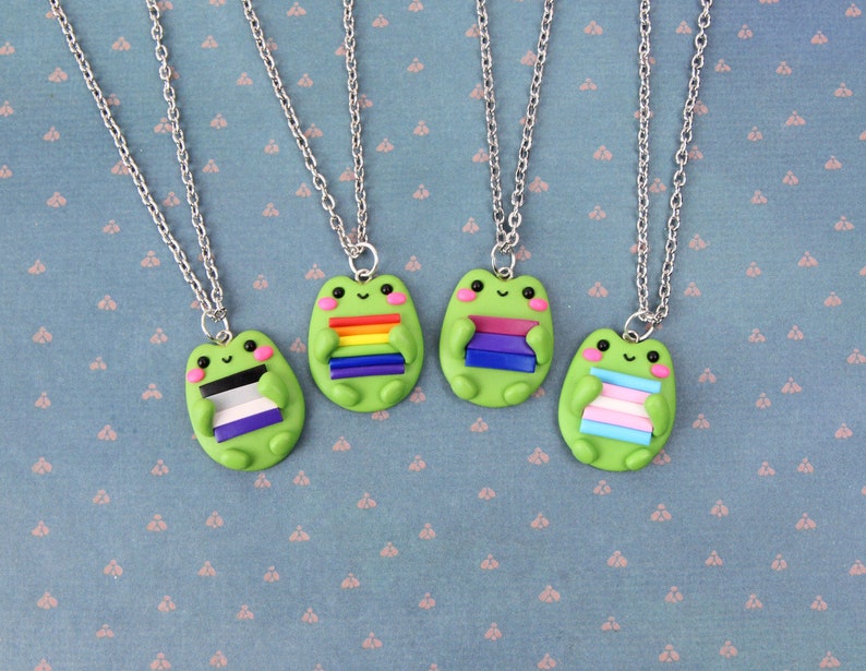 Puede incluir: Cuatro collares de plata con dijes de rana verde que sostienen diferentes banderas del orgullo. Las banderas son arco&iacute;ris, bisexual, asexual y transg&eacute;nero.