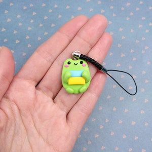 May include: Green frog charm with a blue, yellow, and white striped scarf, holding a yellow and green book. The charm is attached to a black phone strap.