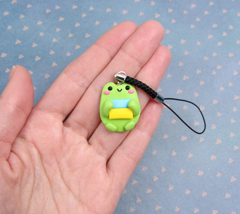 May include: Green frog charm with a blue, yellow, and white striped scarf, holding a yellow and green book. The charm is attached to a black phone strap.