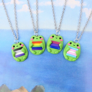 Puede incluir: Cuatro collares de plata con dijes de rana verde que sostienen diferentes banderas del orgullo. Las banderas son arco&iacute;ris, bisexual, asexual y transg&eacute;nero.
