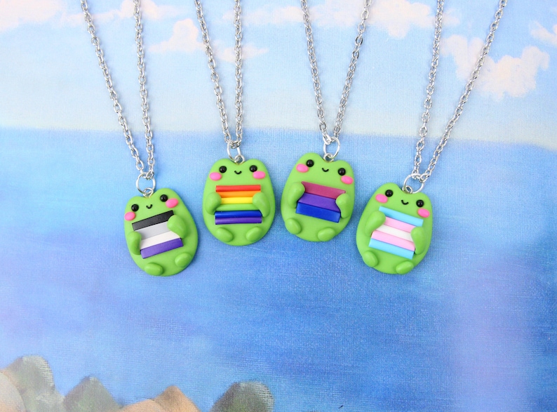 Puede incluir: Cuatro collares de plata con dijes de rana verde que sostienen diferentes banderas del orgullo. Las banderas son arco&iacute;ris, bisexual, asexual y transg&eacute;nero.