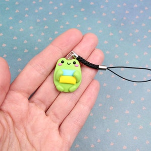 May include: Green frog charm with a blue and yellow stripe, holding a yellow rectangle, attached to a black phone strap.