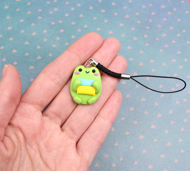 May include: Green frog charm with a blue and yellow stripe, holding a yellow rectangle, attached to a black phone strap.