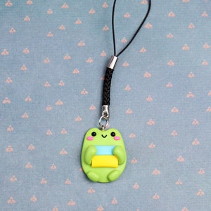 May include: A green frog charm with a yellow, blue, and white striped design, attached to a black phone charm string.