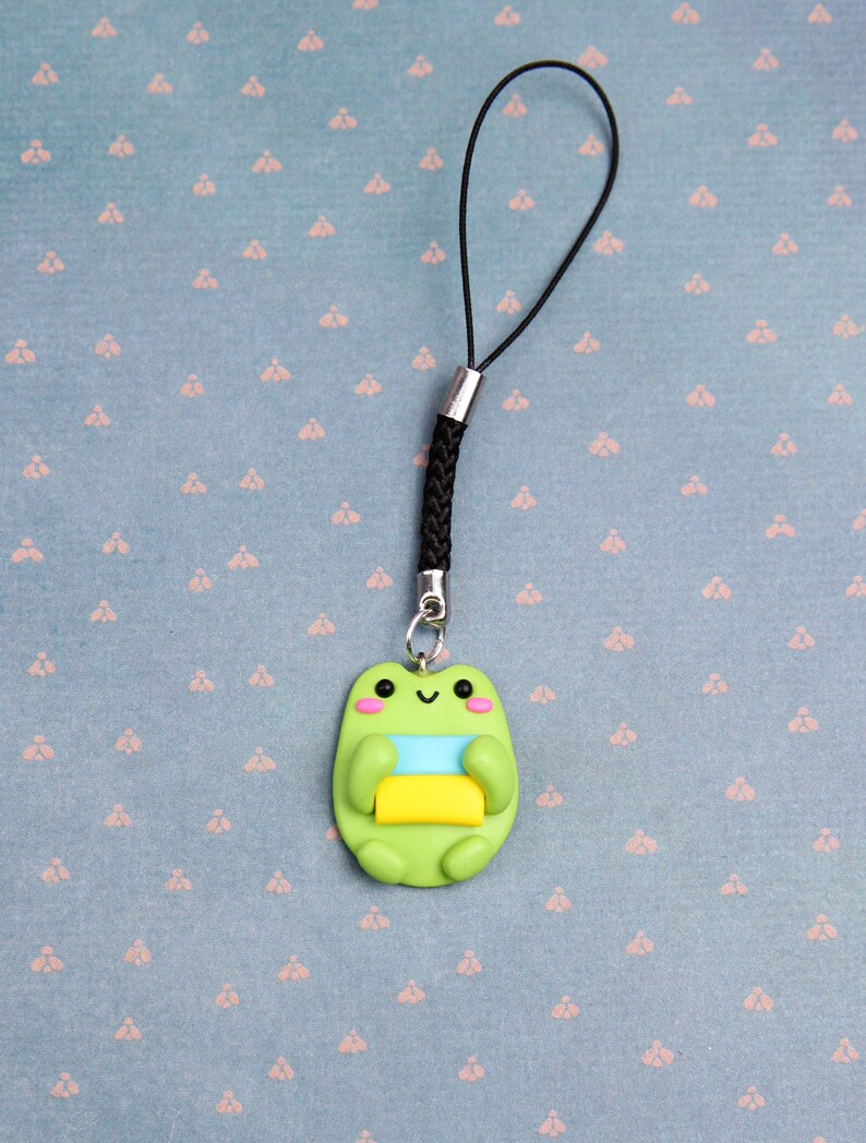 May include: A green frog charm with a yellow, blue, and white striped design, attached to a black phone charm string.