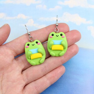 May include: A pair of green frog earrings with silver hooks. Each frog is holding a stack of three colorful squares, blue, light green, and yellow.