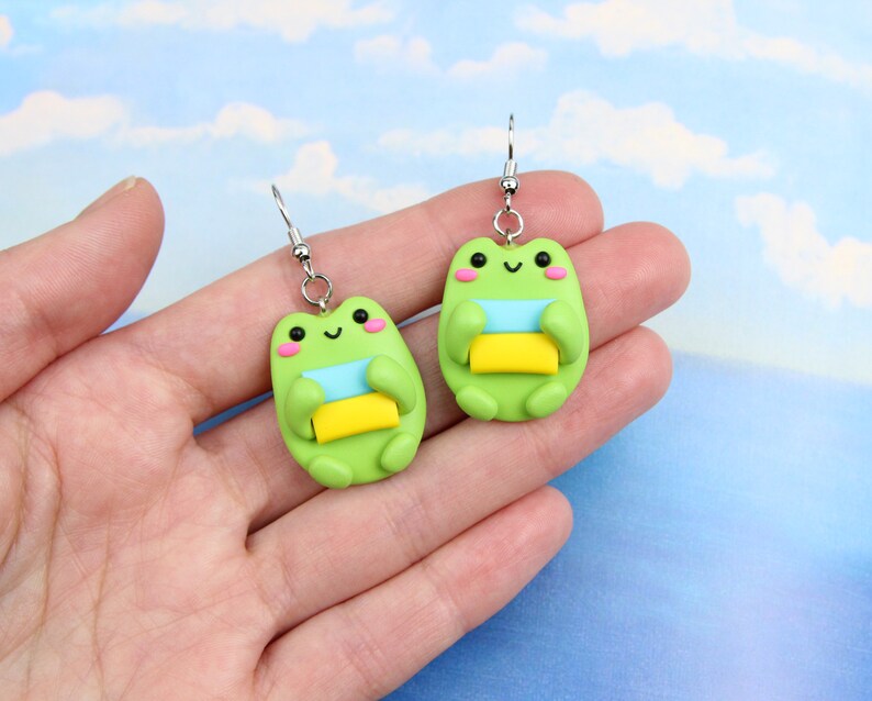 May include: A pair of green frog earrings with silver hooks. Each frog is holding a stack of three colorful squares, blue, light green, and yellow.