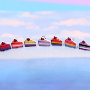 May include: Seven pairs of small, colorful, cake-shaped earrings with a cherry on top. The earrings are arranged in a row on a light blue surface.
